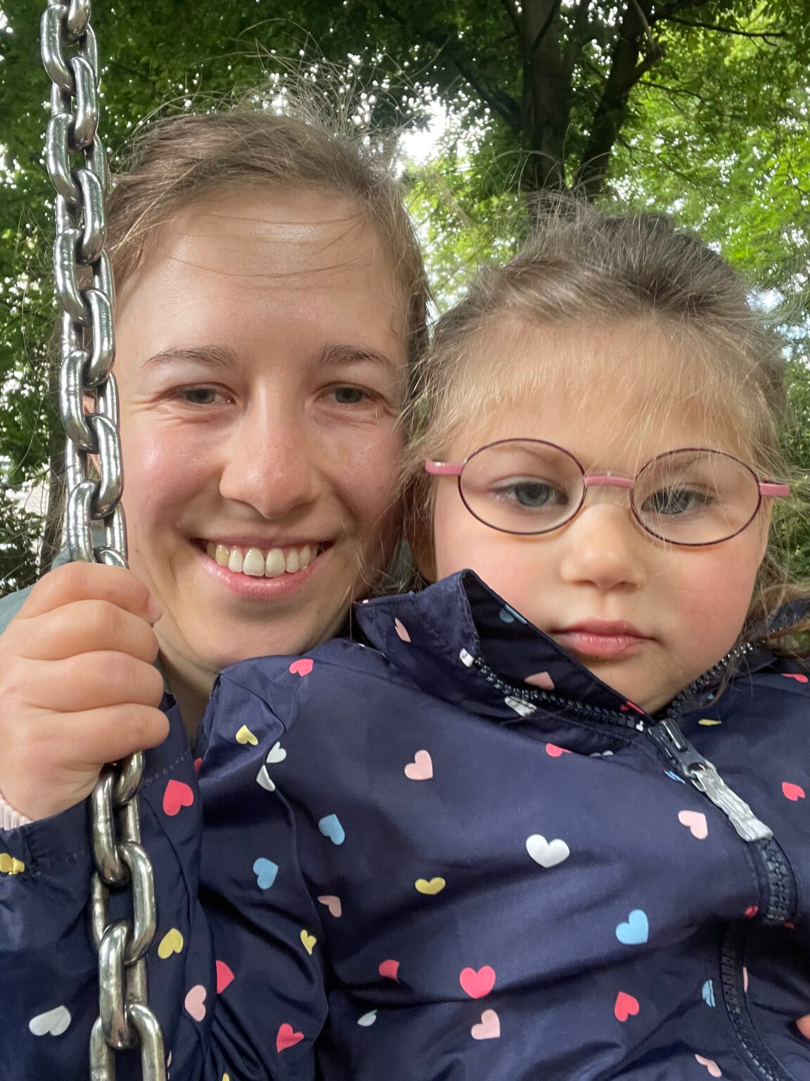 Frau macht ein Selfie mit einem Mädchen, das auf ihrem Schoß auf einer Schaukel sitzt. Beide tragen Regenjacken, das Mädchen blickt ruhig in die Kamera, während die Frau fröhlich lächelt.