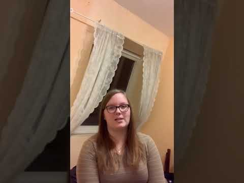 Eine junge Frau mit Brille sitzt vor einem Fenster mit weißen Vorhängen in einem Zimmer mit pastellfarbenen Wänden. Sie blickt ruhig in die Kamera, als würde sie sprechen oder interviewt werden.