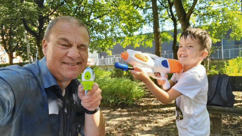 Älterer Mann und ein Junge spielen lachend mit Wasserpistolen im Park. Der Mann kneift ein Auge zu, während der Junge zielt. Beide sind nass und haben sichtlich Spaß.