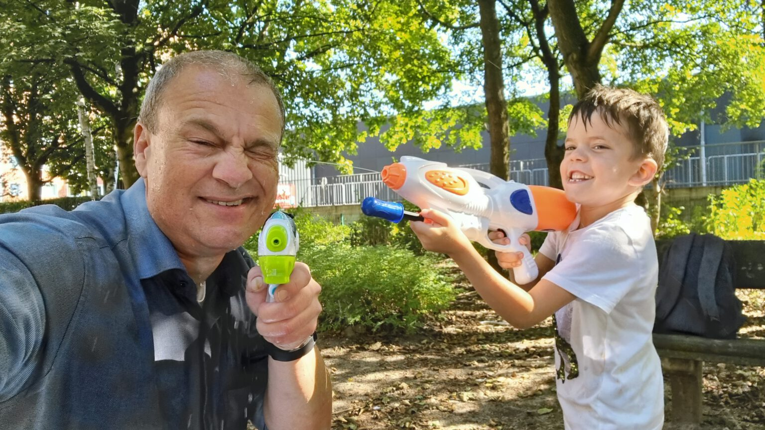 Älterer Mann und ein Junge spielen lachend mit Wasserpistolen im Park. Der Mann kneift ein Auge zu, während der Junge zielt. Beide sind nass und haben sichtlich Spaß.