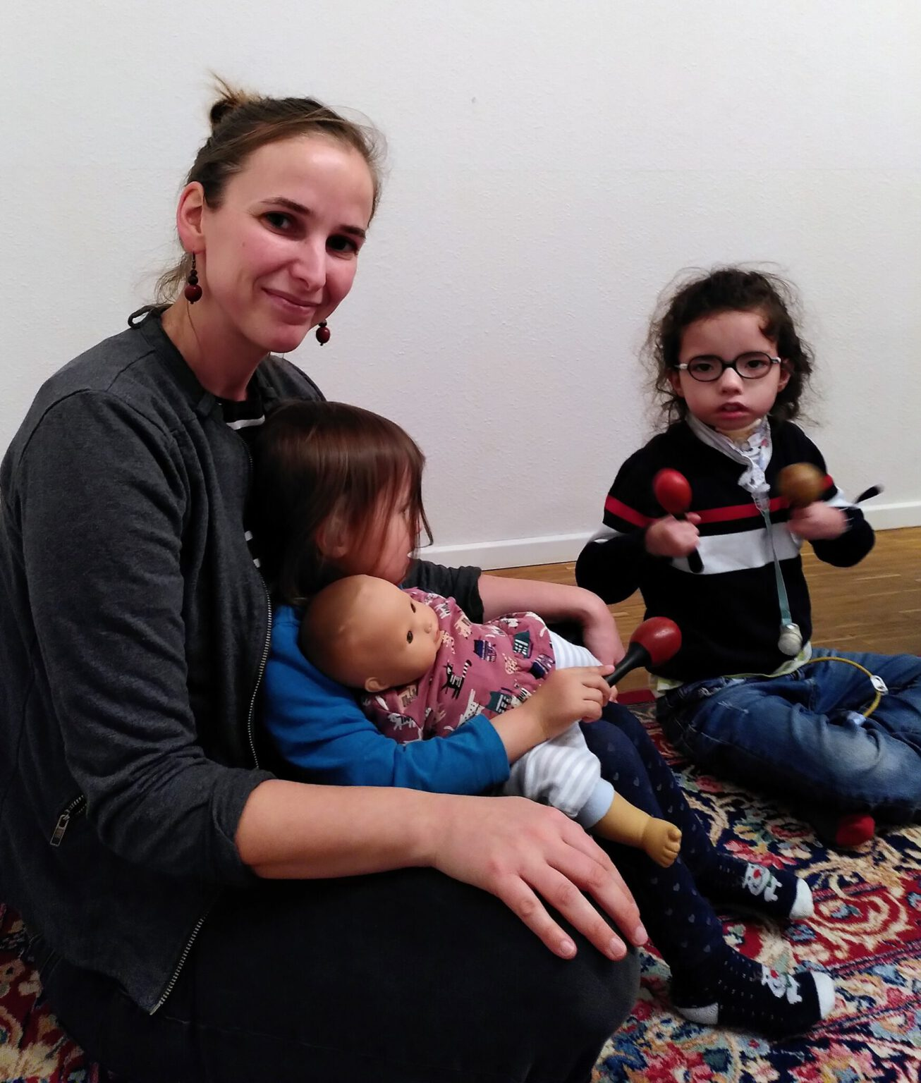 Frau sitzt mit zwei kleinen Kindern auf einem bunten Teppich. Eines der Kinder sitzt auf ihrem Schoß mit einer Puppe im Arm, das andere Kind hält Rasseln und trägt eine Brille sowie ein medizinisches Beatmungsgerät.
