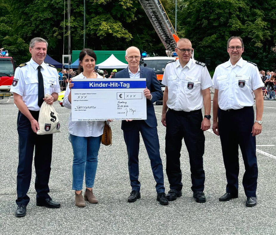 Fünf Personen, darunter vier Männer in Polizeiuniform und eine Frau in Jeans und weißem Hemd, halten gemeinsam einen großen Spendenscheck mit der Aufschrift „Kinder-Hit-Tag“ in der Hand. Im Hintergrund stehen Einsatzfahrzeuge und Zuschauer.