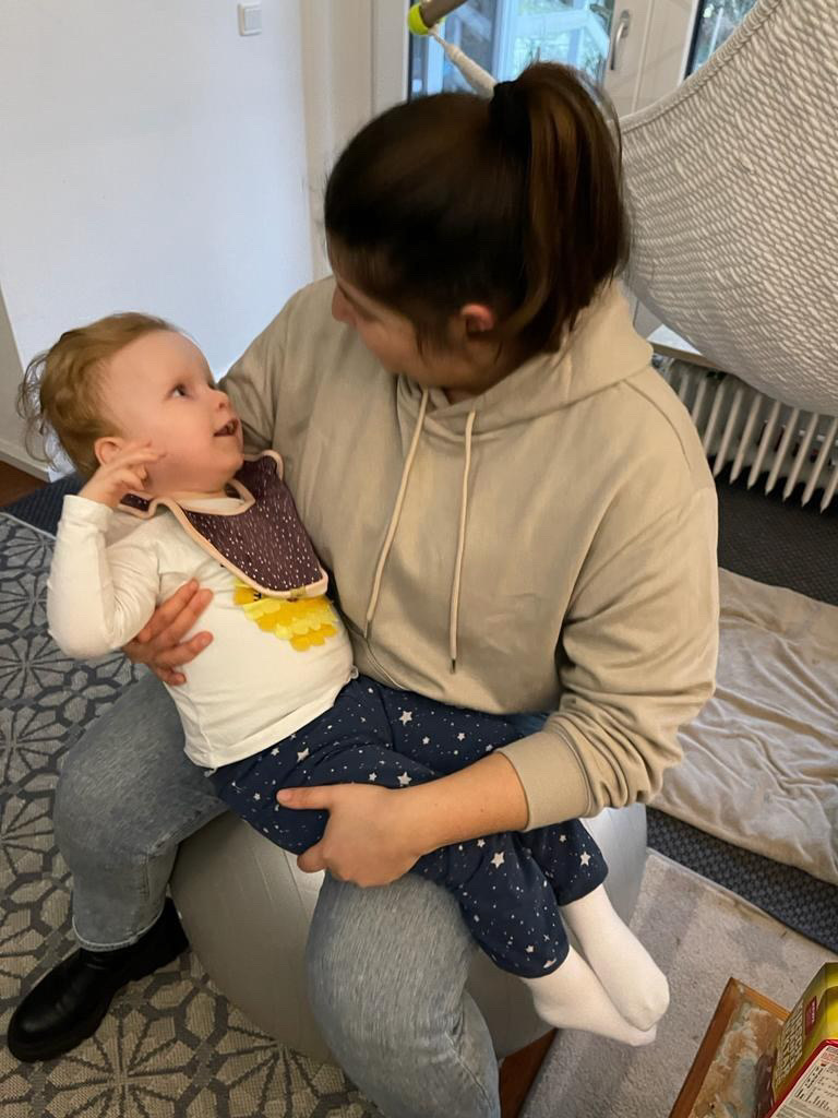 Frau sitzt auf einem Gymnastikball in einem Wohnzimmer und hält ein kleines Mädchen im Arm, das sie liebevoll anschaut. Beide wirken entspannt und verbunden.