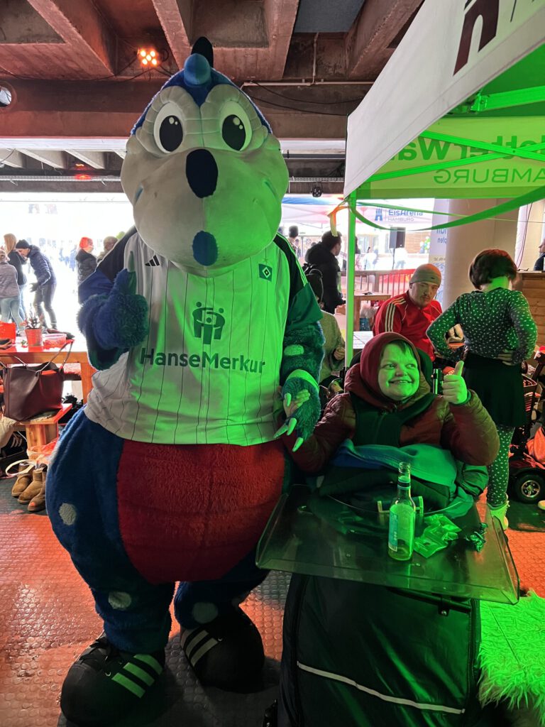 Ein Kind im Rollstuhl gibt lächelnd einen Daumen hoch neben dem HSV-Maskottchen Dino Hermann, das in grünem Licht leuchtet. Die Szene spielt sich unter einem Zelt bei einer Veranstaltung ab.