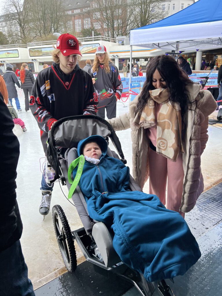 Ein Junge liegt in einem Spezial-Kinderwagen, eingehüllt in eine blaue Decke, begleitet von einer Frau und einem Eishockeyspieler mit rotem Cap auf einer Eislauffläche. Im Hintergrund weitere Personen auf dem Eis.