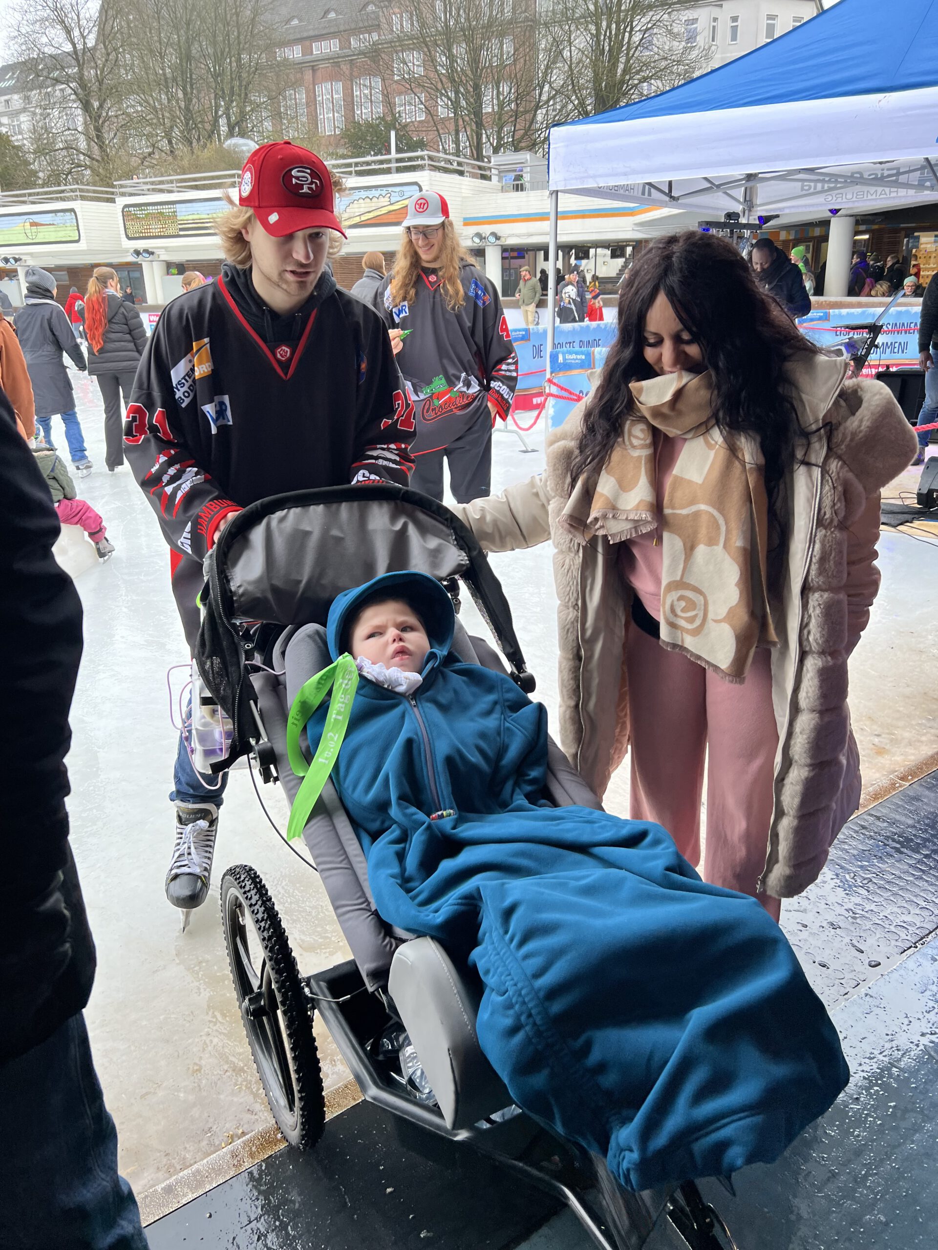 Ein Junge liegt in einem Spezial-Kinderwagen, eingehüllt in eine blaue Decke, begleitet von einer Frau und einem Eishockeyspieler mit rotem Cap auf einer Eislauffläche. Im Hintergrund weitere Personen auf dem Eis.