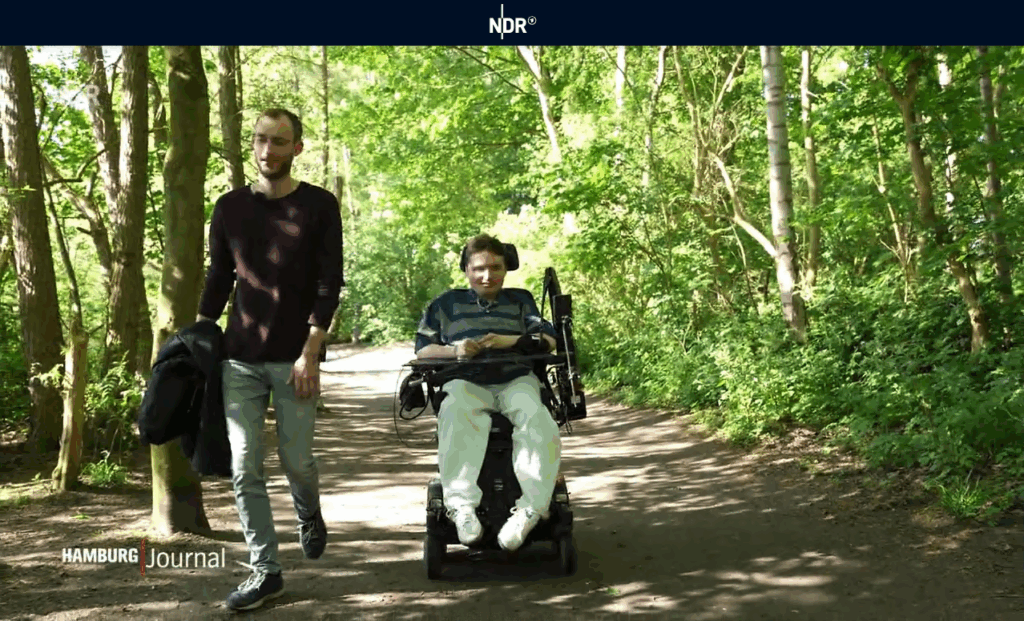 Zwei junge Männer machen einen Spaziergang durch ein Waldstück. Einer läuft, der andere fährt im elektrischen Rollstuhl. Oben im Bild ist das Logo des „Hamburg Journal“ vom NDR zu sehen.