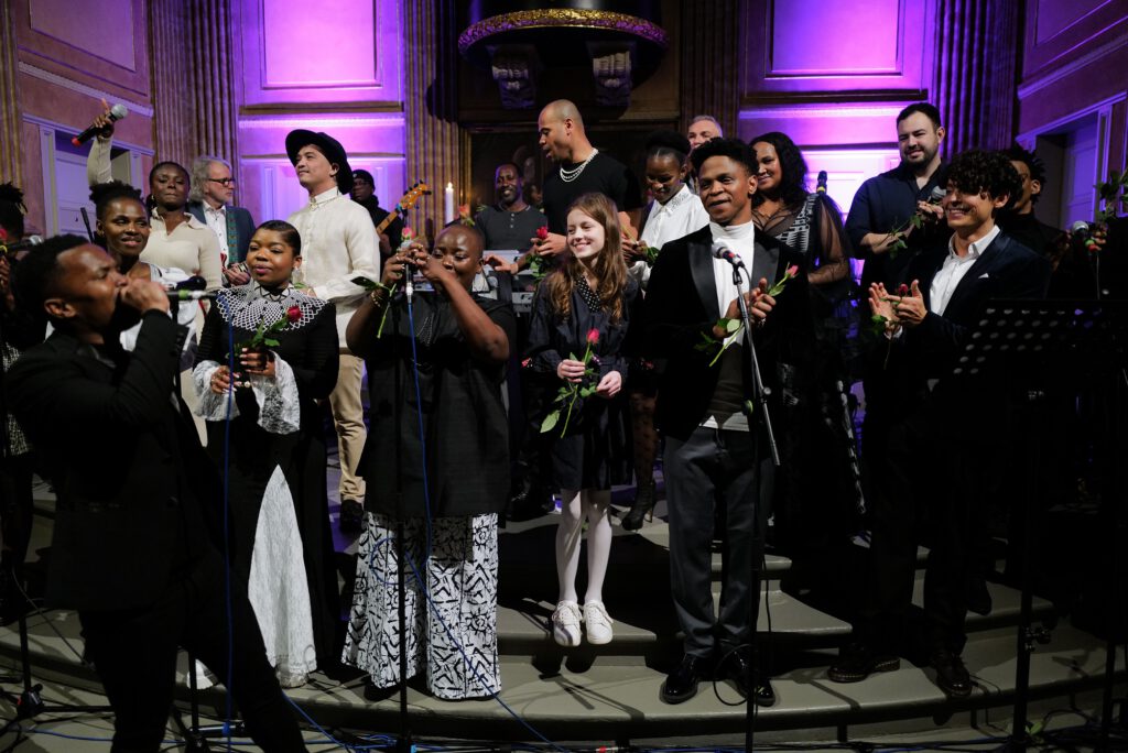 Große Gruppe von Sänger:innen und Musiker:innen steht auf einer festlich beleuchteten Bühne, viele halten Rosen in den Händen und lächeln ins Publikum.